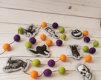May include: A Halloween garland featuring felt balls in orange, purple, and lime green, interspersed with grey felt cutouts. The cutouts display black ink illustrations of a crow, a potion bottle, a jack-o'-lantern, a cat, a tombstone, and a skull.