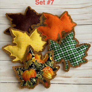 May include: A collection of five fabric leaf-shaped ornaments labeled Set #7. The leaves feature various patterns including solid dark brown, orange textured, yellow grid, green and white plaid, and a black print with orange pumpkins.