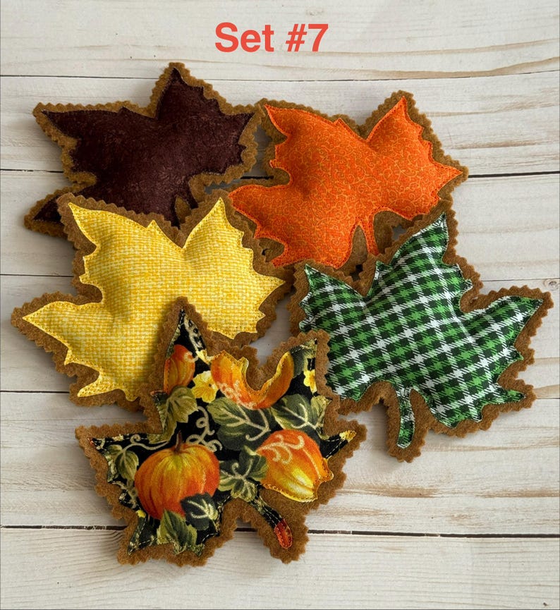 May include: A collection of five fabric leaf-shaped ornaments labeled Set #7. The leaves feature various patterns including solid dark brown, orange textured, yellow grid, green and white plaid, and a black print with orange pumpkins.