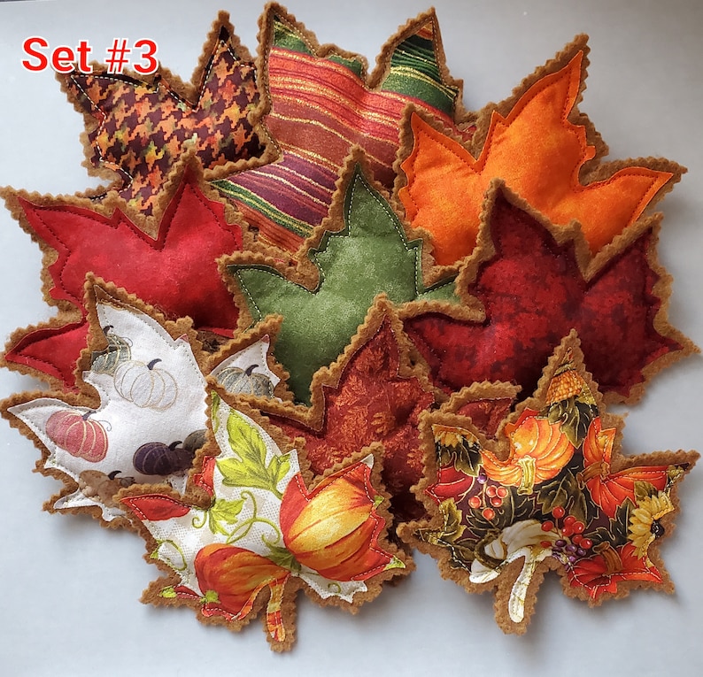 May include: A collection of fabric leaf-shaped ornaments labeled Set #3. The leaves feature various patterns including pumpkins, houndstooth, stripes, and solid autumn colors like orange, red, and green, all finished with brown scalloped edges.