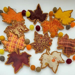 May include: A garland featuring fabric maple leaves in various autumn colors and patterns, including orange, yellow, burgundy, and cream, interspersed with small felt balls in shades of yellow, orange, burgundy, and gray on a thin string.