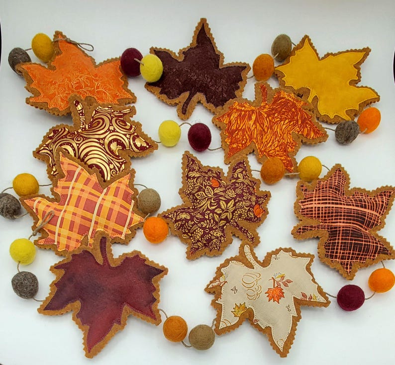 May include: A garland featuring fabric maple leaves in various autumn colors and patterns, including orange, yellow, burgundy, and cream, interspersed with small felt balls in shades of yellow, orange, burgundy, and gray on a thin string.