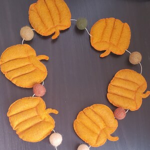 May include: A decorative garland featuring five orange felt pumpkins with stitched details, interspersed with six small felt balls in shades of olive green, tan, brown, and cream, all strung together on a thin white cord in a circular arrangement.