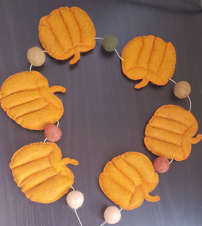 May include: A decorative garland featuring five orange felt pumpkins with stitched details, interspersed with six small felt balls in shades of olive green, tan, brown, and cream, all strung together on a thin white cord in a circular arrangement.
