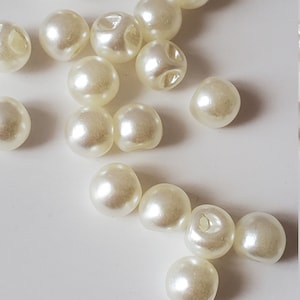 May include: A collection of off-white, round beads with a pearlescent sheen. Each bead has a small hole, suggesting they are for stringing. The beads are scattered on a white surface, creating a simple, elegant aesthetic.