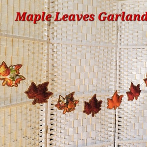 May include: A Maple Leaves Garland featuring fabric maple leaf cutouts in various autumn colors including green, orange, red, and brown, some with pumpkin and floral patterns, strung together on a thin cord against a white woven background.