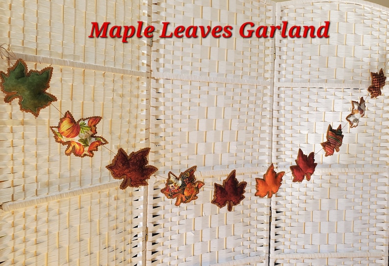 May include: A Maple Leaves Garland featuring fabric maple leaf cutouts in various autumn colors including green, orange, red, and brown, some with pumpkin and floral patterns, strung together on a thin cord against a white woven background.