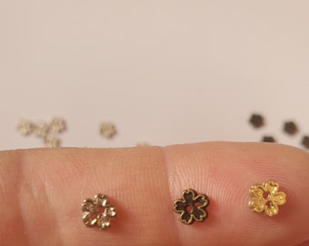 May include: Three small metal flower-shaped beads or buttons, each with a central hole. The left bead is silver-toned, the middle bead is dark bronze, and the right bead is gold-toned. These tiny floral components are arranged in a horizontal row.