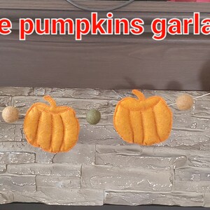 May include: A garland featuring orange felt pumpkins and small round felt balls in beige, green, and muted red colors strung along a thin cord. The text "Large pumpkins garland" is printed in white with a red outline at the top of the image.