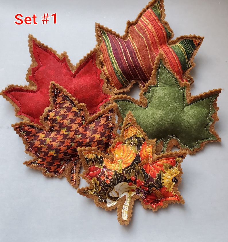 May include: A set of five fabric maple leaf ornaments labeled Set #1. The leaves feature various patterns including solid red, green, striped, houndstooth, and a pumpkin print. Each leaf has a brown felt backing with decorative pinked edges.