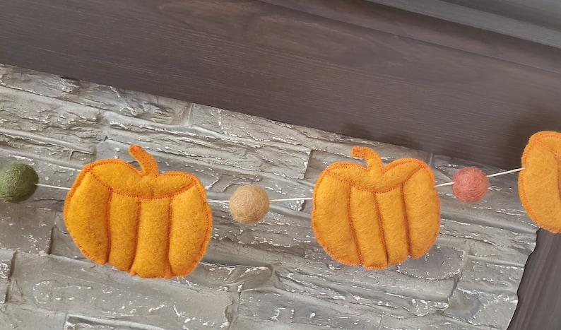 May include: A garland featuring orange felt pumpkins and small felt balls in olive green, tan, and muted red, all strung together on a thin white cord. The pumpkins have visible stitched detailing and a soft, textured appearance throughout the piece.