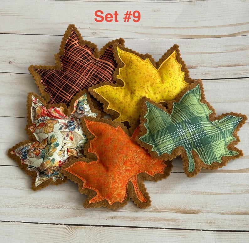 May include: A set of five fabric leaf-shaped ornaments labeled Set #9. The leaves feature various patterns including a black and orange grid, solid yellow, green plaid, orange floral, and a multi-colored autumn floral print on a cream background.