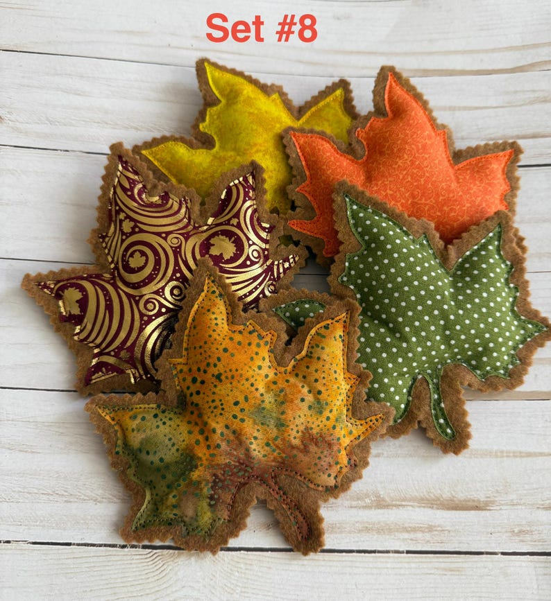May include: A collection of five fabric leaf-shaped ornaments labeled Set #8. The leaves feature various patterns including gold swirls on burgundy, green with white polka dots, solid orange, solid yellow, and a mottled yellow-green-orange print.