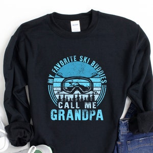 May include: Black sweatshirt with a blue and white graphic design. The graphic features a pair of ski goggles, a mountain, and the text "My Favourite Ski Buddies Call Me Grandpa".