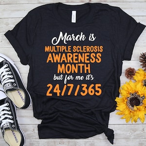 May include: Black t-shirt with the text "March is Multiple Sclerosis Awareness Month but for me it's 24/7/365" in white and orange lettering.