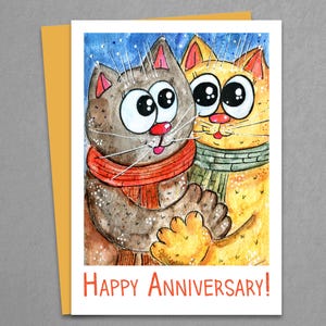 Op de afbeelding: Een kaart met een aquarel-illustratie van twee katten met grote ogen, die sjaals dragen en elkaar omhelzen. De tekst "Happy Anniversary!" is in oranje onderaan de kaart gedrukt.