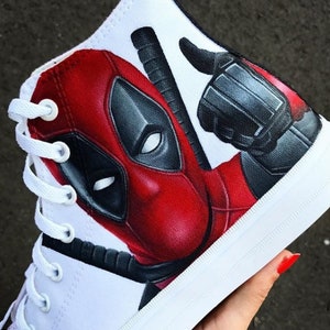 May include: White high-top canvas shoes with a hand-painted design of Deadpool's face and a raised fist. The design is in red, black, and white.