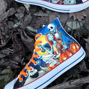 May include: A pair of white high-top sneakers with orange laces, hand-painted with characters from the Nightmare Before Christmas. The design features Jack Skellington and Sally, along with other characters from the film, including Zero the ghost dog. The sneakers are set against a backdrop of a starry night sky and pumpkins.