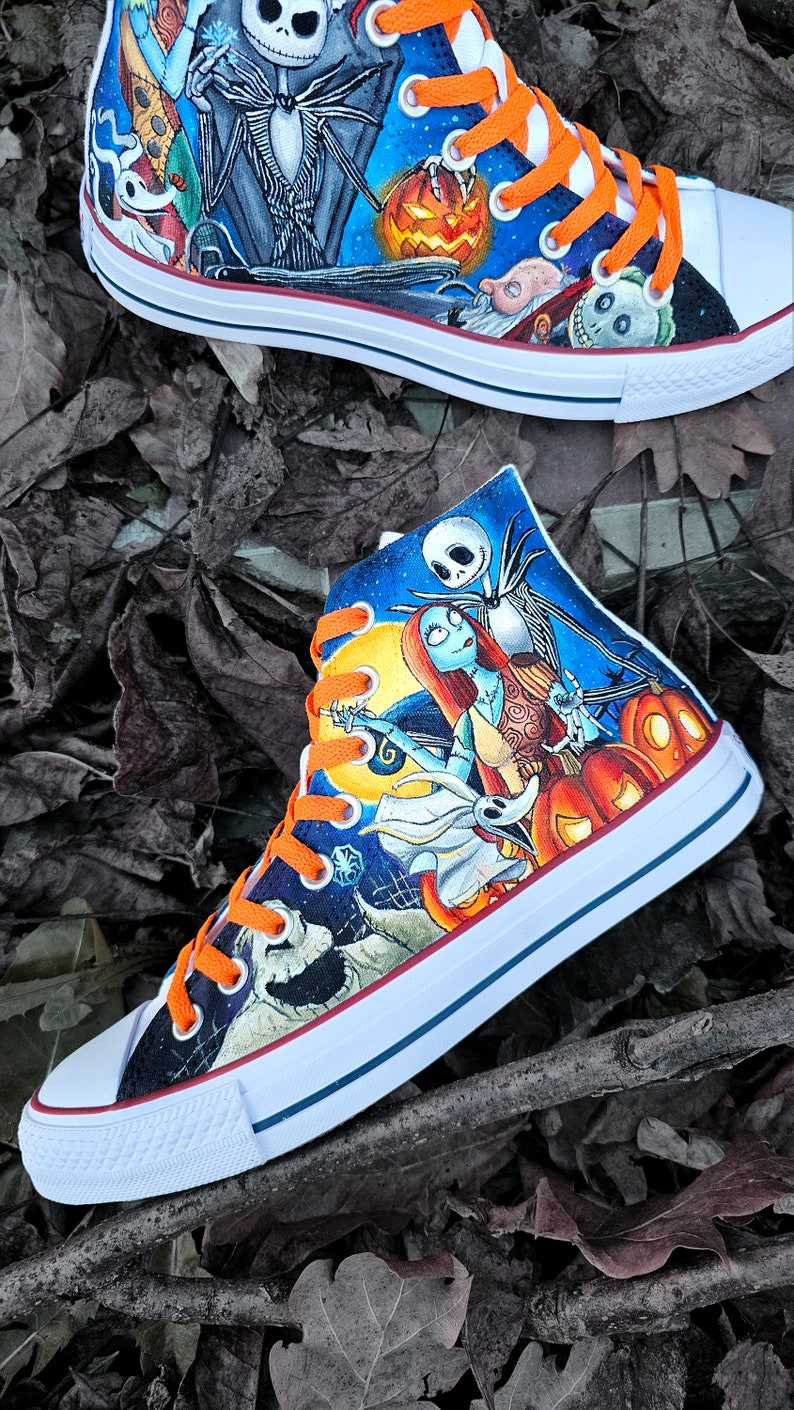 May include: A pair of white high-top sneakers with orange laces, hand-painted with characters from the Nightmare Before Christmas. The design features Jack Skellington and Sally, along with other characters from the film, including Zero the ghost dog. The sneakers are set against a backdrop of a starry night sky and pumpkins.