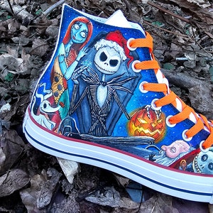 May include: A pair of high-top sneakers with a hand-painted design featuring characters from the Nightmare Before Christmas. The design includes Jack Skellington, Sally, and Zero. The sneakers have orange laces and a white sole.