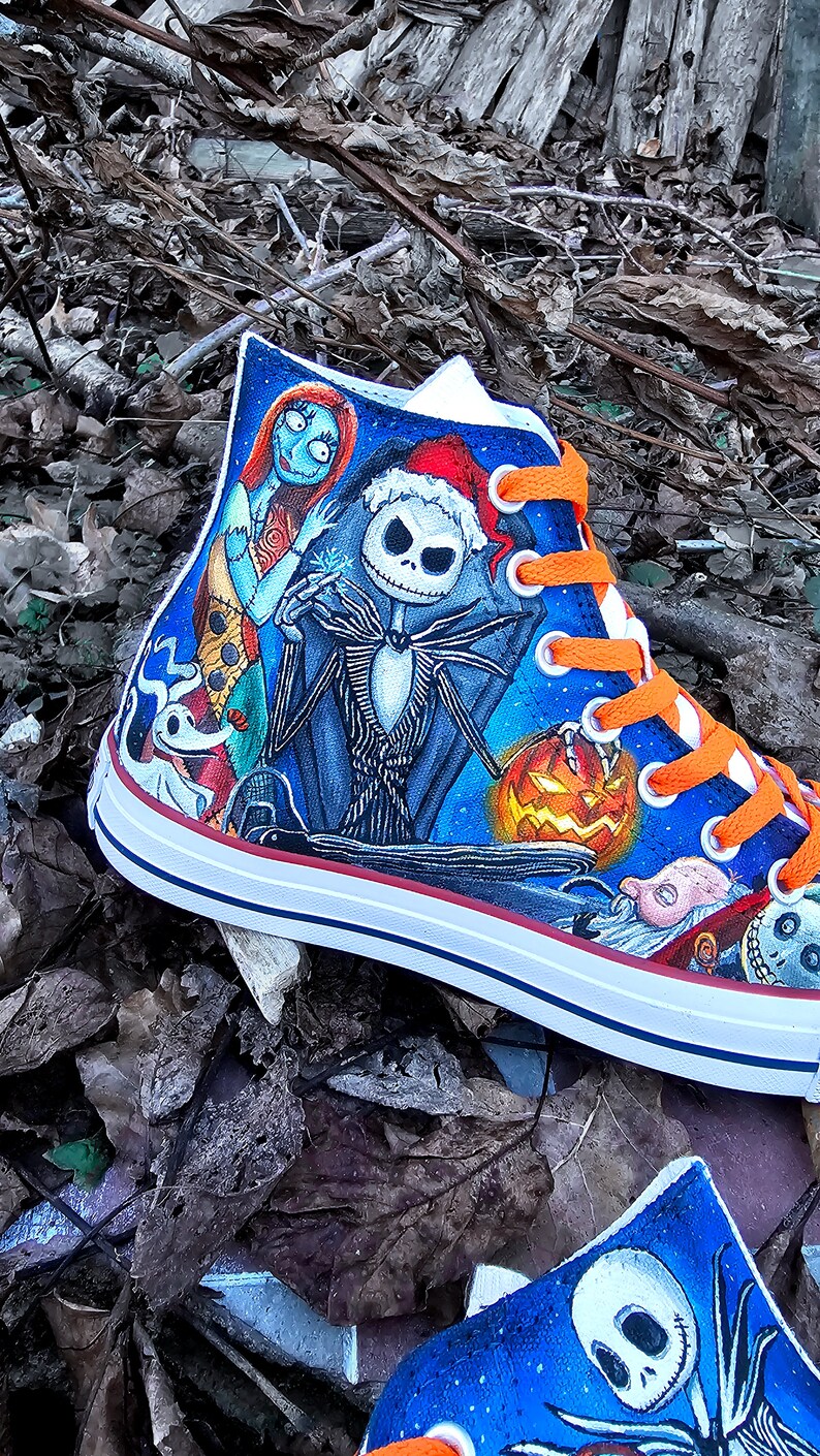 May include: A pair of high-top sneakers with a hand-painted design featuring characters from the Nightmare Before Christmas. The design includes Jack Skellington, Sally, and Zero. The sneakers have orange laces and a white sole.