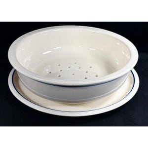 May include: A round, off-white ceramic berry bowl with a matching saucer. The bowl features a series of drainage holes at the bottom and a thin, dark blue horizontal stripe encircling the exterior of both the bowl and the saucer base.