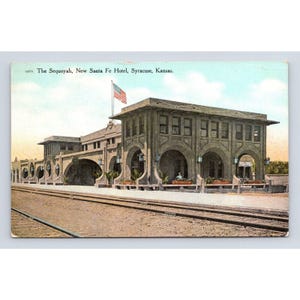 May include: A vintage postcard showing The Sequoyah, New Santa Fe Hotel, Syracuse, Kansas. The building features a gray stone facade with multiple arched openings, a flat roof, and an American flag flying from a pole on the roof above the tracks.