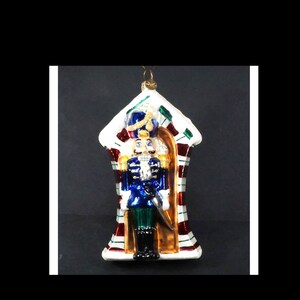 May include: A glass Christmas ornament featuring a nutcracker standing in a candy cane striped house. The nutcracker is wearing a blue and gold uniform with a red and white hat. The ornament is white with red and green stripes.