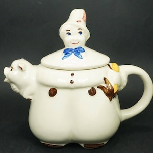 May include: White ceramic teapot shaped like a person wearing a hat and holding a pig. The teapot has brown spots and a blue bow.