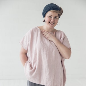 Könnte beinhalten: Eine Frau trägt ein locker sitzendes, rosa Leinenoberteil mit kurzen Ärmeln. Sie trägt außerdem graue Leinenhosen. Sie lächelt und trägt einen dunkelblauen Kopftuch.