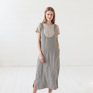 May include: A woman wearing a gray linen overall dress with a light brown linen top underneath. She is standing on a white wood floor and has her hands in her pockets.