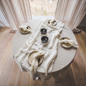 Könnte beinhalten: Ein runder Tisch, der zum Essen gedeckt ist, bedeckt mit einer beigen Tischdecke und einem weißen Tischläufer. Der Tisch ist mit Tellern, Servietten und kleinen Schalen gedeckt. Der Hintergrund umfasst ein Fenster mit transparenten Vorhängen und Blick auf Bäume.