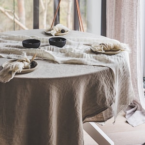 May include: A round, light brown linen tablecloth with a white linen runner on top. There are four place settings with black bowls and white napkins. A brown tripod floor lamp with a white lampshade is in the background.