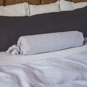May include: A bed with a dark gray body pillow and a light gray bolster pillow. The pillows are on a white bedspread with a gray duvet. The headboard is a warm brown color.