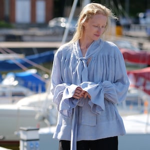 May include: A woman wearing a light blue linen blouse with a bow tie and black shorts. She is standing on a dock with boats in the background.