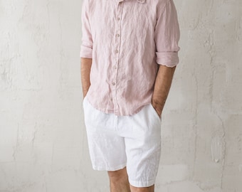 May include: A man wearing a light pink linen shirt and white linen shorts. He is standing in front of a white wall.