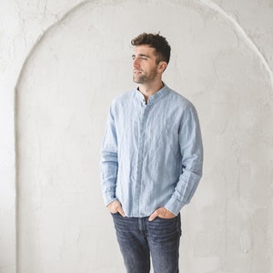 Op de afbeelding: Een man draagt een lichtblauw linnen overhemd met een mandarijnkraag en donkerblauwe jeans. Hij staat voor een witte muur met een boog.