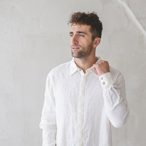 May include: A man wearing a white linen shirt with a button-down collar and long sleeves. He is standing in front of a white wall and has his hand on his collar.