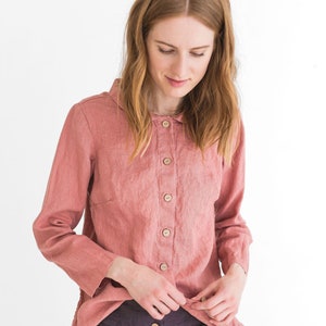 May include: A woman wearing a pink linen shirt with a collar and button fastening. She is also wearing dark purple linen trousers with a button fastening.