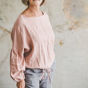 Könnte beinhalten: Eine Frau trägt ein hellrosa Leinenoberteil mit langen Ärmeln und einer gerafften Taille. Sie trägt auch graue Leinenhosen.