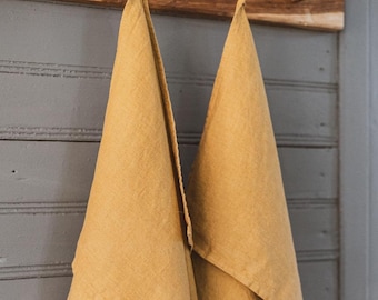 May include: Two mustard-yellow linen towels hanging from a rustic wooden rack. The towels are folded and draped, showcasing their textured fabric. The rack is mounted on a gray, paneled wall, creating a simple, farmhouse aesthetic.