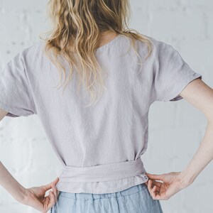 以下が含まれることがあります： 薄い紫色のリネン素材のトップスに、同色のリボンベルトを合わせた女性。薄いブルーのリネン素材のパンツを履いています。