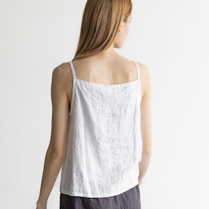 May include: A white linen tank top with thin straps and a relaxed fit. The model is wearing a dark gray linen skirt.
