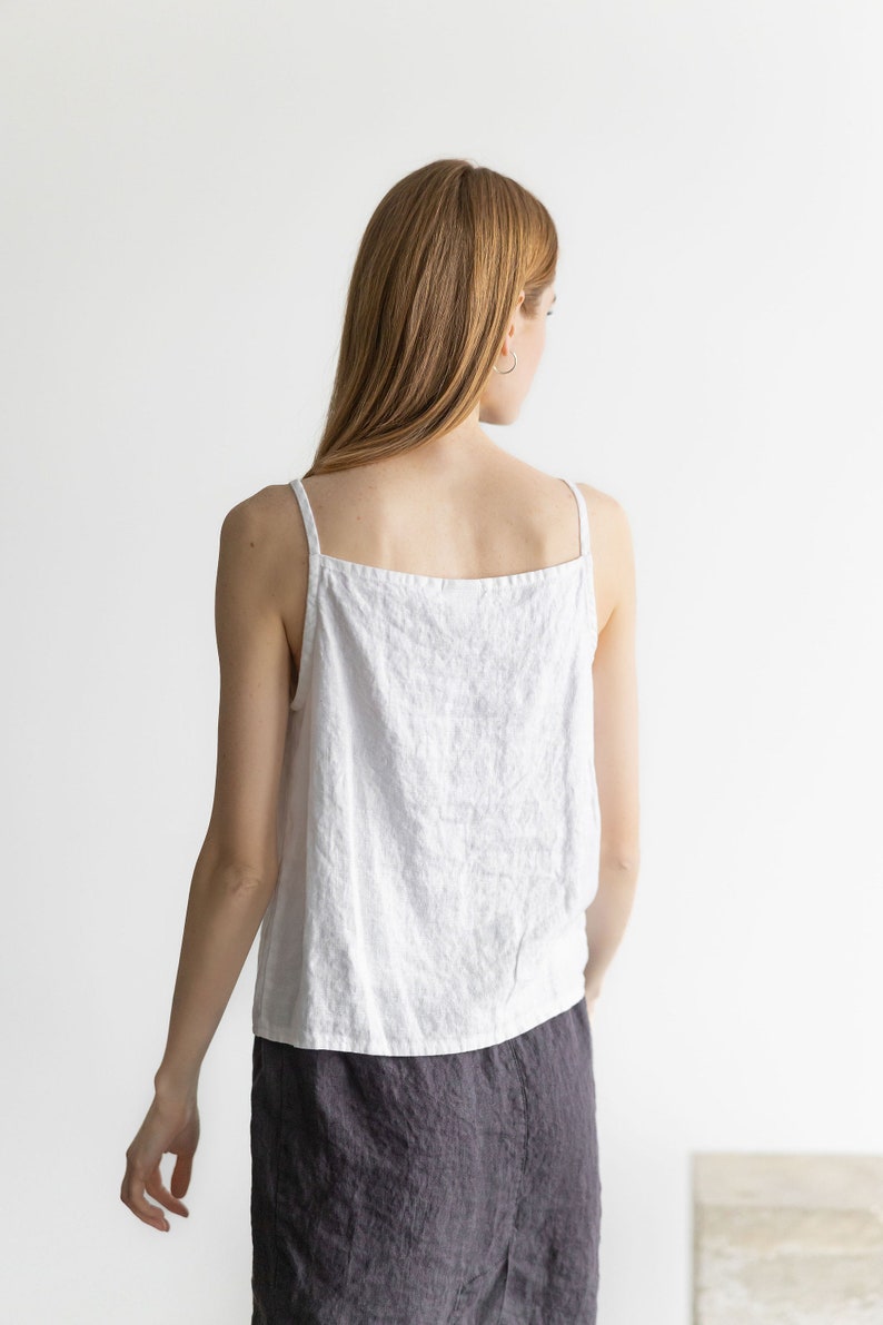 May include: A white linen tank top with thin straps and a relaxed fit. The model is wearing a dark gray linen skirt.