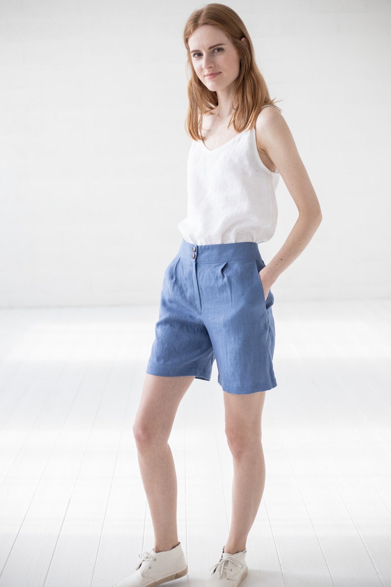May include: A woman wearing a white linen tank top and blue linen shorts. She is standing in a white room with her hands in her pockets.
