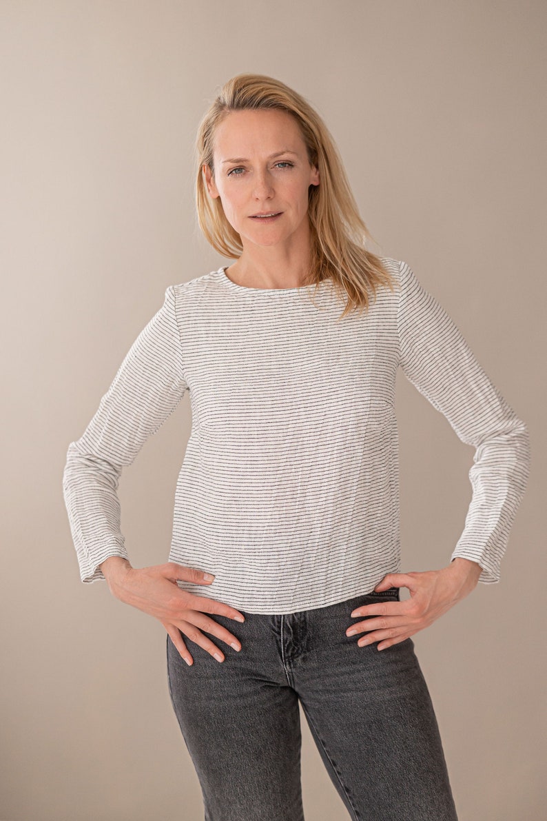 May include: A long-sleeved top featuring a white base with thin, horizontal black stripes, a round neckline, and a straight hemline, paired with dark gray denim jeans. The garment has a textured fabric appearance and a fitted silhouette.