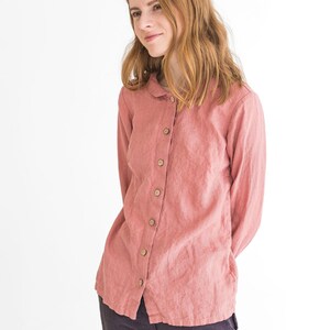 May include: A woman wearing a long-sleeved, pink linen shirt with a button-up front and a collar. She is wearing dark grey trousers.