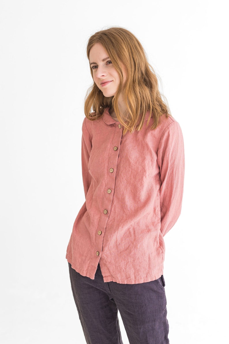 May include: A woman wearing a long-sleeved, pink linen shirt with a button-up front and a collar. She is wearing dark grey trousers.