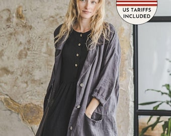 May include: A charcoal grey long-sleeved coat featuring a notched collar, four front buttons, and two large patch pockets, worn over a black button-down dress. A circular badge in the upper right corner displays the US flag and text US TARIFFS INCLUDED.