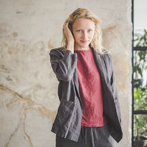 May include: A woman wearing a gray linen blazer over a red linen top and matching gray linen pants. She is standing in front of a textured wall.